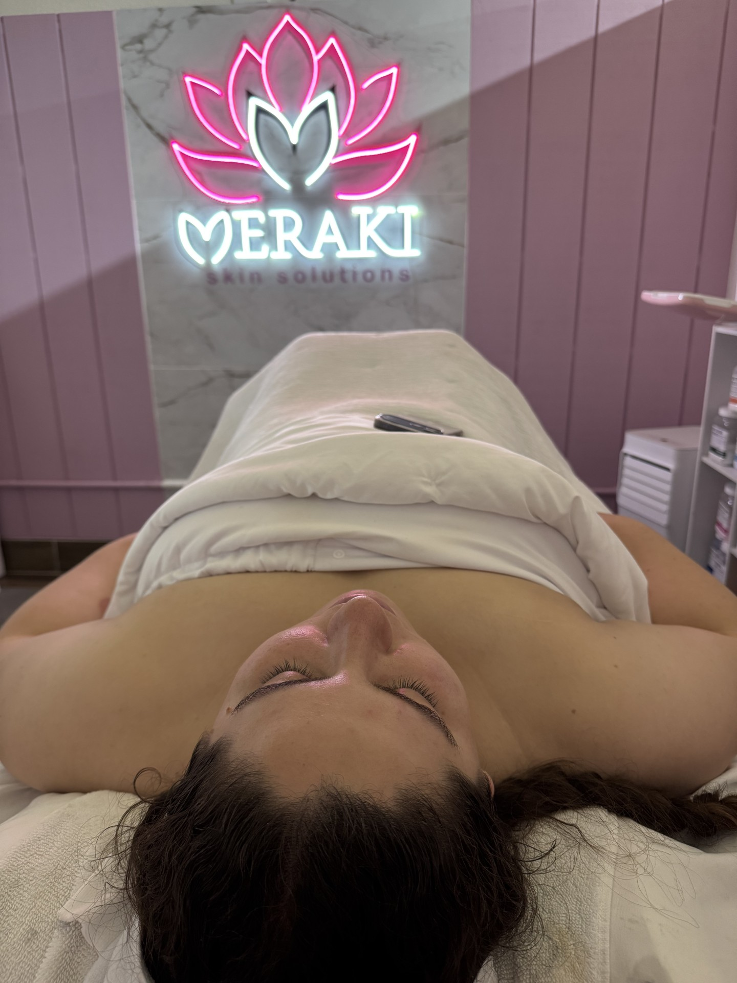 Client relaxing during a facial at Meraki Skin Solutions, under the neon studio sign